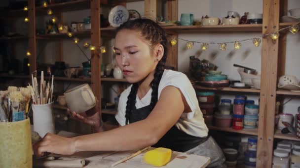 Young woman potter smoothing out the clay cup using a wet brush — 图库视频影像