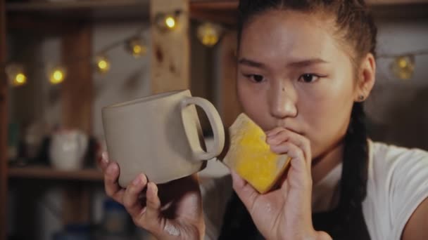 Young woman potter smoothing the surface of cup handle using a sponge — Vídeo de Stock