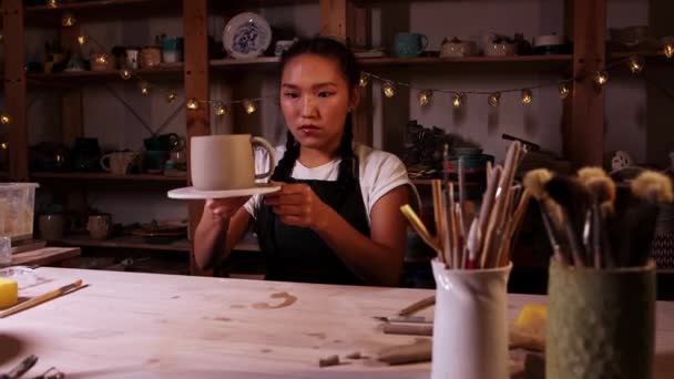Кераміка в студії - азіатська гончарка пробує на ручці до чашки і дивиться на неї — стокове відео