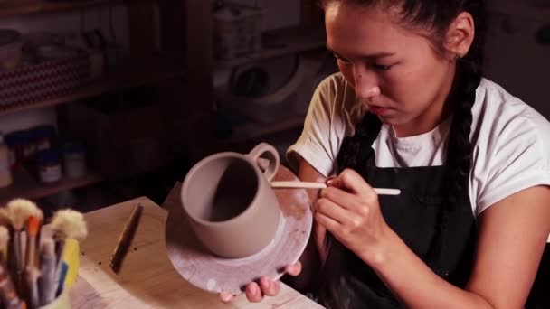 Pottery in the art studio - asian woman holding a cup on the plate and smearing clay on the joint on the handle and a cup using a tool — Αρχείο Βίντεο