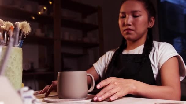 Pottery in the art studio - woman potter puts a piece of clay on the joint between the handle and cup - brushes on the foreground — Vídeos de Stock