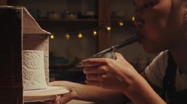 Pottery in the studio - young woman potter manually sprays white powder paint on the clay cup by blowing in the device