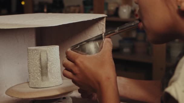 Pottery in the studio - young woman manually sprays white powder paint on the clay cup by blowing in the device — Vídeo de stock