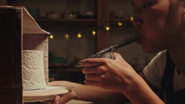 Pottery in the studio - young woman potter manually sprays white powder paint on the clay cup by blowing in the device — Vídeo de stock