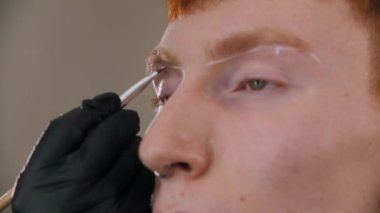 Kaş ustası kızıl erkek müşterisinin kaşlarına kılavuz çizgiler çiziyor.