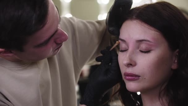 スタジオで彼のかわいい女性のクライアント上の眉の下部にワックスを適用男化粧アーティスト — ストック動画