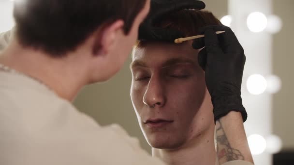 Um mestre sobrancelha homem aplicando cera nos cabelos da sobrancelha de seu cliente masculino — Vídeo de Stock