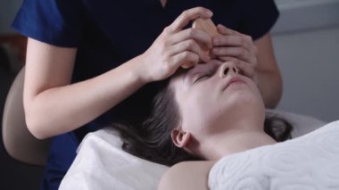 Kadın masajcı, kadın müşterisinin alnında tahta bir aletle derisini yüzüyor.