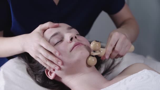 Mujer masajista usando un rodillo de madera debajo de la mandíbula de su cliente femenino — Vídeo de stock