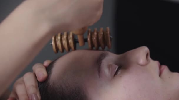Masajista usando una herramienta de rodillo acanalado de madera en la frente de su cliente femenino — Vídeo de stock