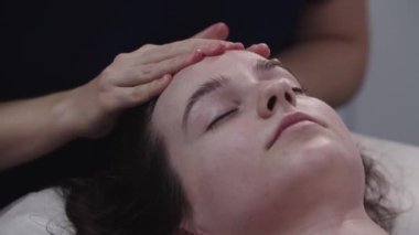 Masajcı kadın müşterisinin derisindeki parmaklarıyla yüz masajı yapıyor. Alnına masaj yapıyor.