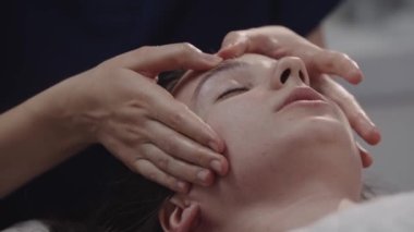 Masajcı, müşterisinin derisindeki parmaklarıyla yüz masajı yapıyor ve alnına masaj yapıyor.