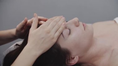 Masajcı, kadın müşterisinin temiz cildindeki parmaklarıyla yüz masajı yapıyor. Alnına masaj yapıyor.