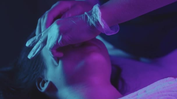 Женщина-массажист делает массаж лица пальцами на своей клиентке - неоновое освещение — стоковое видео