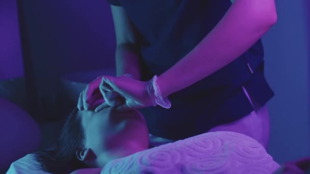 Rubia joven masajista haciendo un masaje facial usando sus dedos en su cliente femenino - iluminación de neón — Vídeos de Stock