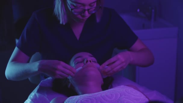 Массажист-блондинка делает массаж лица с помощью нефрита на своей клиентке - неоновое освещение — стоковое видео