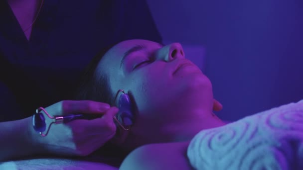 Женщина-массажист делает массаж лица с помощью нефритового ролика на своей клиентке - темно-фиолетовое неоновое освещение — стоковое видео