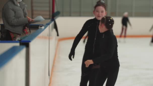Two little girls figure skaters meeting near the sideboard of public rink and talking — Stock Video