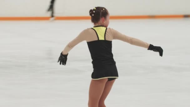 Маленькая девочка фигуристка в тренировочном черно-желтом костюме катается на общественном катке и падает — стоковое видео