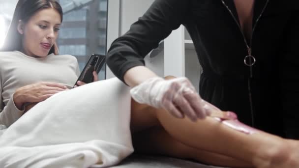Восковая процедура - нанесение розового горячего воска на ногу с помощью лопатки, в то время как женщина сидит на диване и смотрит на свой телефон — стоковое видео