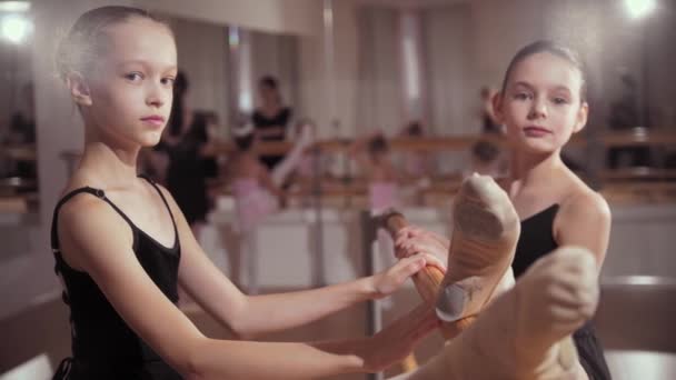 Entrenamiento de ballet - grupo de pequeñas bailarinas entrenando junto al espejo y dos chicas estirándose junto al estrado y mirando a la cámara — Vídeos de Stock