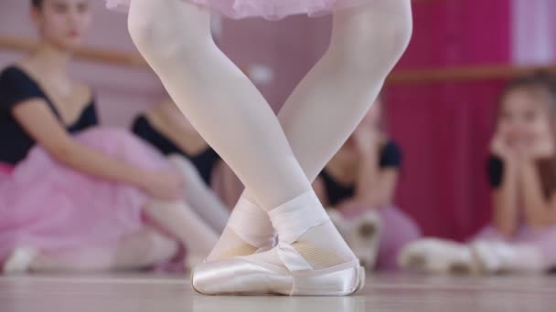 Балетная подготовка - девушки-балерины в красивых платьях, сидящие на полу, и одна из них демонстрирует балетные позиции на переднем плане — стоковое видео
