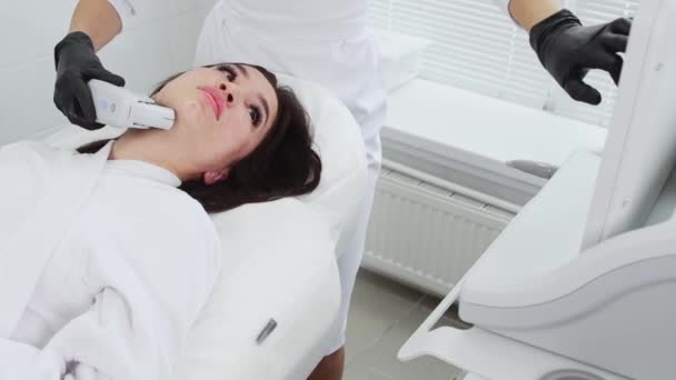 Masaje corporal - joven morena sometida a un procedimiento de masaje eléctrico en la cara - trabajando debajo de la barbilla y mirando al monitor — Vídeos de Stock