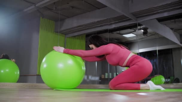 Mujer joven en traje rosa haciendo ejercicios en el gimnasio apoyándose en la pelota de fitness y estirando su columna vertebral — Vídeos de Stock