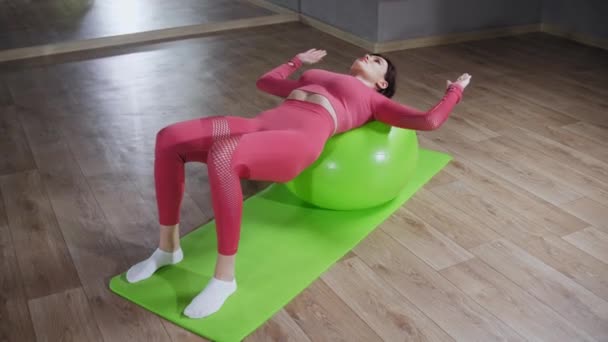 Femeie tânără în costum roz sprijinindu-se pe mingea de fitness și făcând exerciții fizice cu mâinile ei — Videoclip de stoc