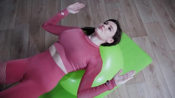 Joven mujer bonita en traje rosa apoyándose en la pelota de fitness y haciendo ejercicio con sus manos — Vídeos de Stock