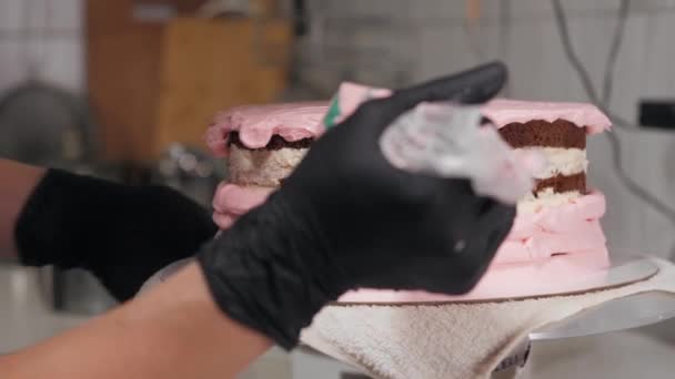 El chef de pastelería aplica crema dulce rosa en los lados de un pastel en blanco — Vídeo de stock