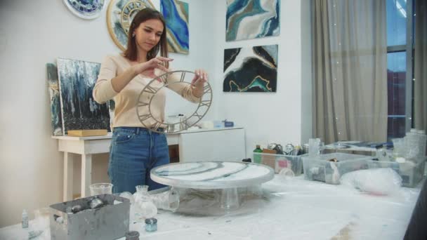 Молодая женщина заканчивает эпоксидную роспись смолой в виде часов в художественной студии — стоковое видео