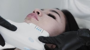 Genç esmer kadın kanepede uzanıyor ve çenesinin altında elektrikli masaj yaptırıyor.