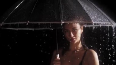 Siyah mayo giymiş genç bir kadın yağmurun altında duruyor ve elinde şemsiye tutuyor. Kenardan su damlıyor.