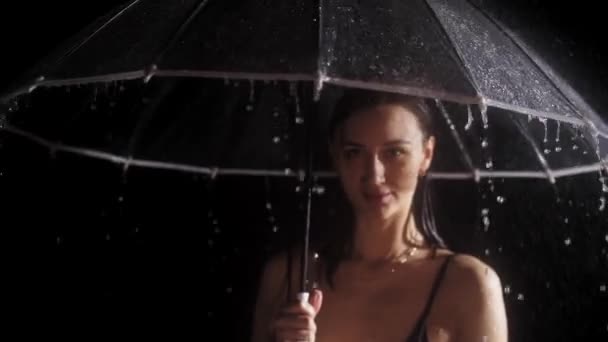 Siyah mayo giymiş genç bir kadın yağmurun altında duruyor. Elinde şemsiye tutuyor ve çeviriyor. — Stok video