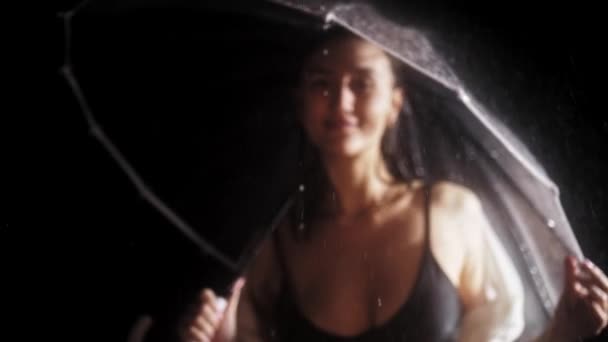 Jovem flerte mulher de fato de banho preto em pé sob a chuva - segurando um guarda-chuva e olha para a câmera — Vídeo de Stock