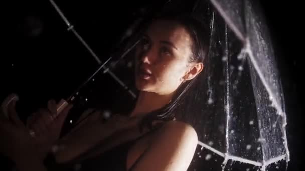 Siyah mayo giymiş, elinde şemsiyeyle yağmurun altında duran genç ve çapkın bir kadın uzaklığa bakıyor. — Stok video