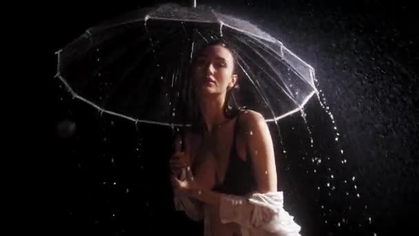 Siyah mayo giymiş genç bir kadın yağmurun altında duruyor. Kameraya bakarken şemsiye tutuyor ve çeviriyor. — Stok video