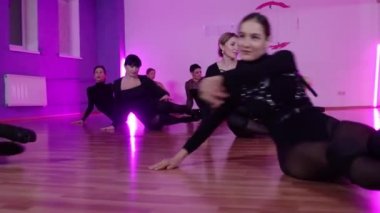 Siyah elbiseli bir grup seksi kadın stüdyoda yerde mor ışıkla dans ediyor.