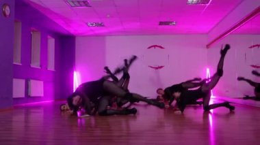 Siyah elbiseli bir grup seksi kadın dansçı senkronize olarak yerden kalkar ve kameraya bakar.