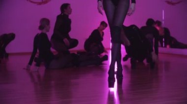 Siyah elbiseli bir grup seksi kadın dans antrenmanına hazırlanıyor ve bir tanesi ön planda dans hareketleri yapıyor.