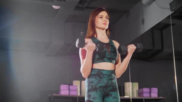 Молодая женщина в спортивном костюме накачивает мышцы рук гантелями — стоковое видео