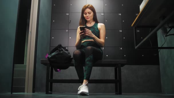 Jovem mulher em trajes esportivos sentada no vestiário de seu ginásio e usando seu telefone — Vídeo de Stock