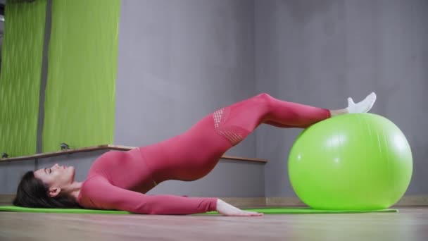 Mladá žena ve sportovním kostýmu opírá nohy o fitness míč a dělá cvičení — Stock video