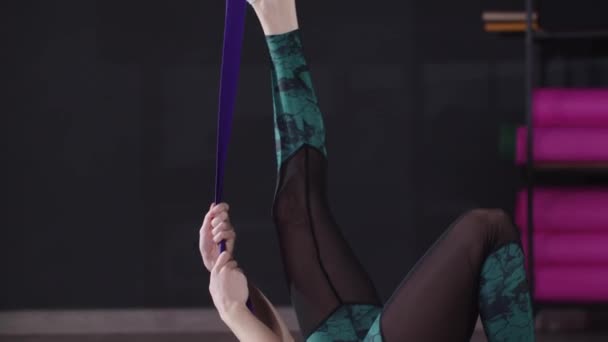 Mujer joven y deportiva que usa una banda elástica en el pie mientras se estira en el gimnasio — Vídeos de Stock