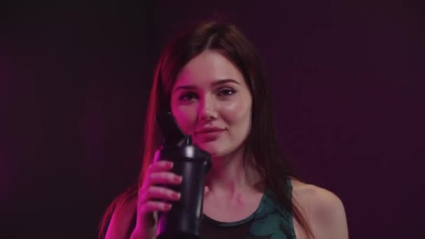 Jonge mooie vrouw in sportieve top kijkt in de camera en drinkt water uit plastic beker in neon verlichting — Stockvideo