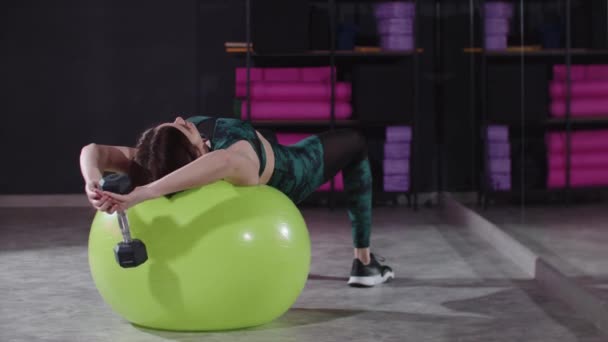 年轻女子躺在健身球上，双手托着哑铃在健身大厅里锻炼 — 图库视频影像