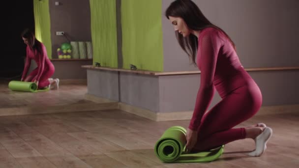 Fitness training - jonge vrouw in roze kostuum de uitrol van de yoga mat en begint een oefening — Stockvideo