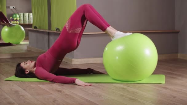 Fitness trénink - mladá žena v růžovém kostýmu se opírá nohama o fitness míč a dělá cvičení s ohýbáním kolena — Stock video