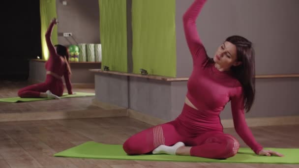 Fitness trénink - mladá žena v růžovém kostýmu sedí na podložce jógy a naklání se do stran — Stock video